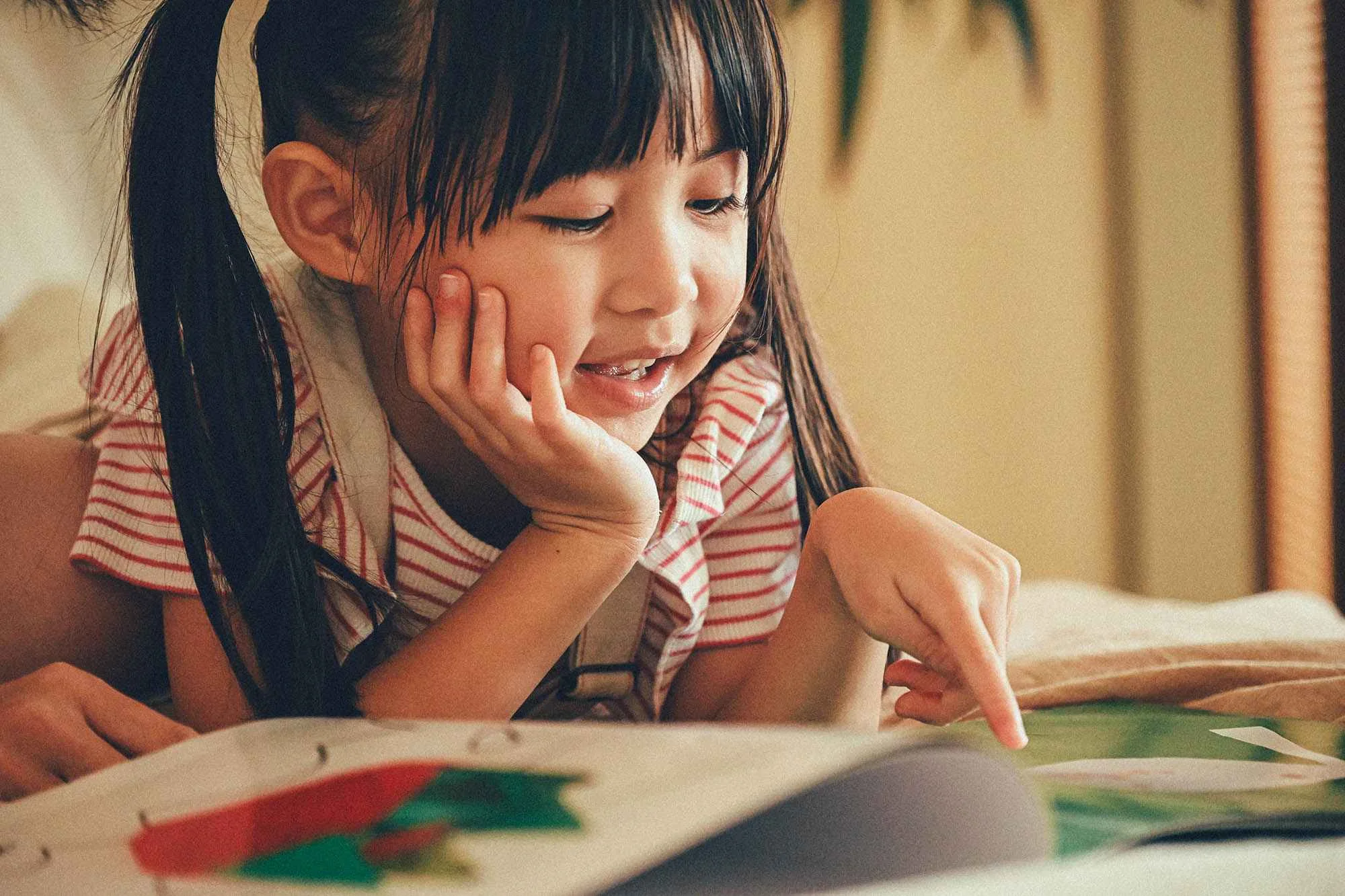 A young girl, around 3 to 5 years old, is lying on her stomach while looking joyfully at a colorful book. Dressed in a red-and-white striped ruffle-sleeve top and white overalls, she rests her chin on one hand and points at a page with the other. Her expression is bright and engaged. The book contains illustrations, possibly of paper crafts or children’s artwork, suggesting a personalized gift or creative keepsake. This warm and candid moment captures the delight and curiosity typical of preschool kids when receiving thoughtful birthday gifts. 一位約3至5歲的小女孩趴在床上，開心地翻閱一本色彩繽紛的書。她穿著紅白條紋的荷葉邊短袖上衣和白色吊帶褲，一手撐著下巴，一手指著書頁，臉上流露出投入與興奮的表情。書中展示的是手工藝或小朋友的畫作，彷彿是一份個人化的藝術禮物。這個溫馨自然的瞬間完美展現了幼稚園小朋友收到精心準備生日禮物時的快樂與好奇。 Birthday Gifts for Kids aged 3-5 years, 香港的3-5歲小朋友生日禮物, Art Classes for Kids aged 3-5, 3-5歲兒童畫班