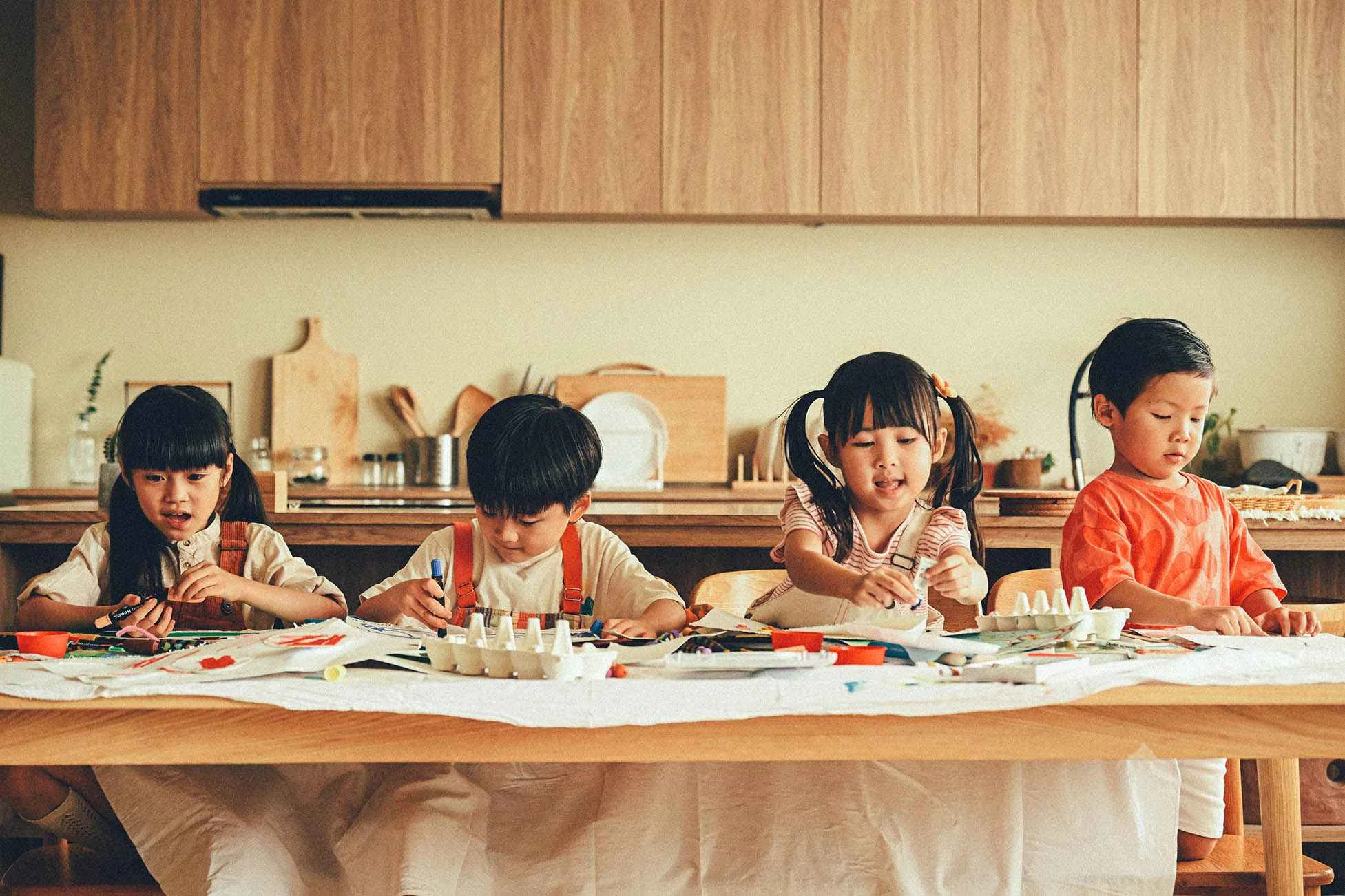 A group of children enthusiastically engaged in painting and drawing activities at a wooden table in a cozy kitchen setting. They are using colorful crayons, markers, and paint palettes on a white tablecloth, surrounded by art supplies. This vibrant scene highlights the benefits of painting for kids, fostering creativity, self-expression, and cognitive development. 一群小朋友在溫馨的廚房裡圍著木桌開心地進行繪畫和塗鴉活動。桌上鋪著白色桌布，擺滿了彩色蠟筆、彩色筆和顏料盤，孩子們全情投入於創作之中。這幅生動的場景展現了小朋友畫畫的好處，有助於培養創造力、自我表達和認知發展。